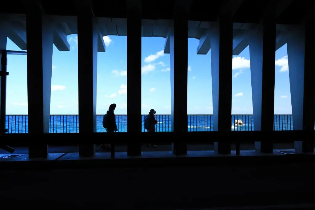 柱が並ぶ屋外通路を歩いている二人のシルエットと、奥に広がる青い海